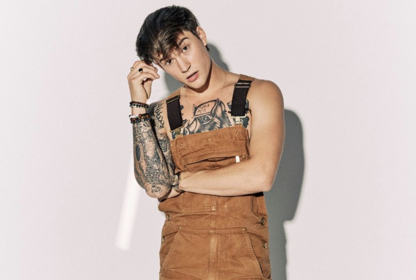Cameron Whitcomb wearing brown dungarees with his arms folded in front of a white backdrop.